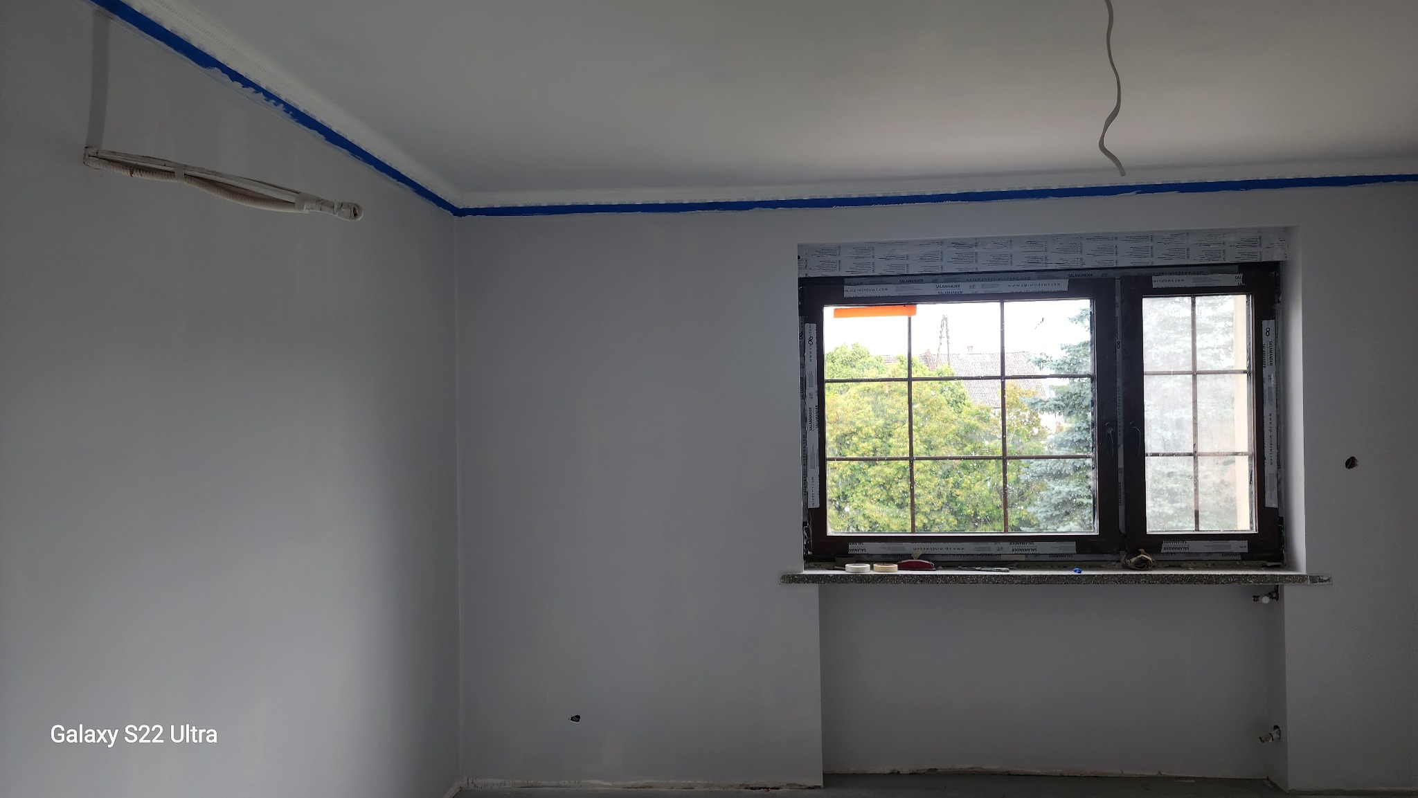 Szare, świeżo pomalowane wnętrze pokoju z oknem. Biały sufit z niebieską taśmą malarską przy listwach. Widoczne przewody i wystające kable.