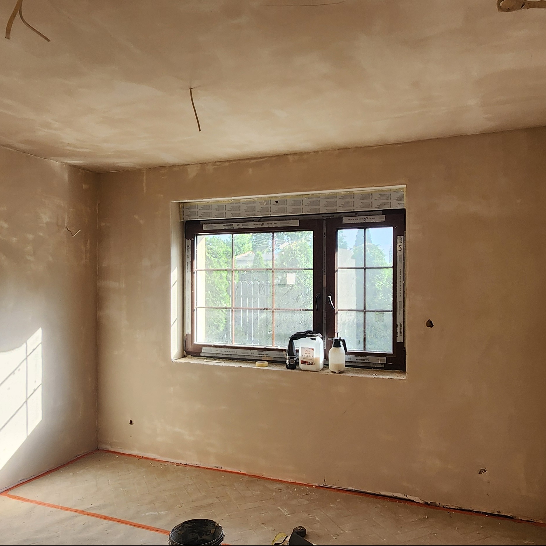 Pokój po tynkowaniu ścian i sufitu, z widocznym oknem i narzędziami na parapecie. Na podłodze widoczne ślady po remoncie.