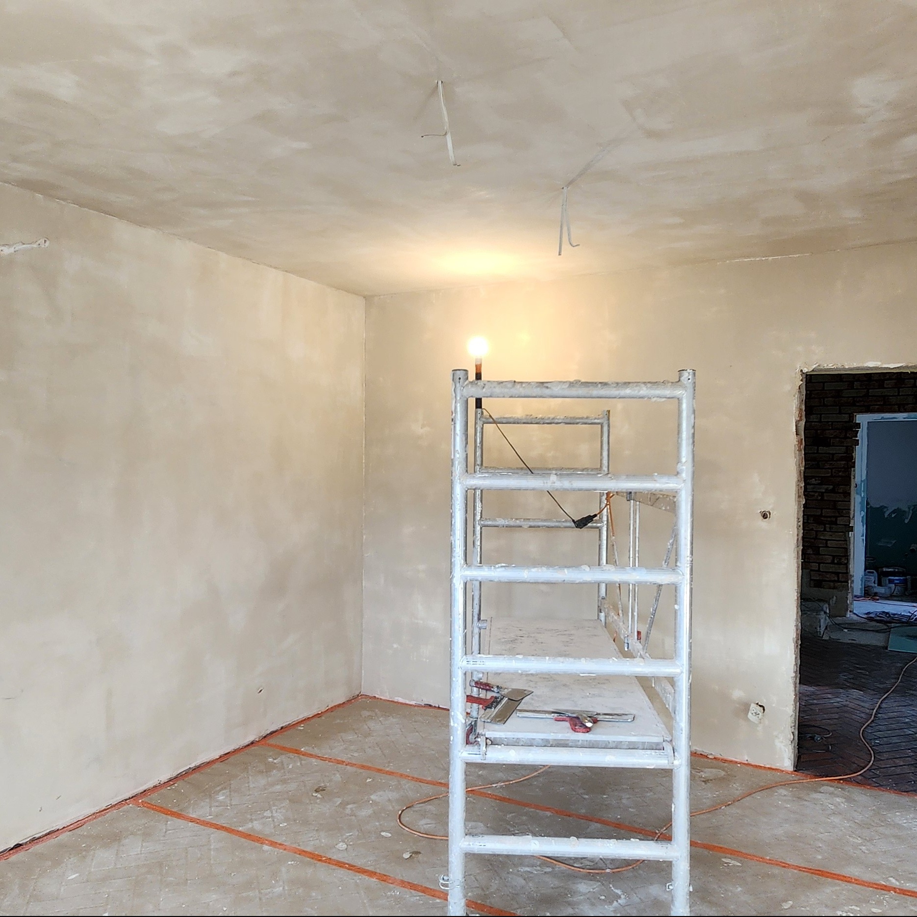 Wnętrze po gładziowaniu ścian i sufitu, widoczne rusztowanie z narzędziami. Na podłodze pomarańczowe taśmy wyznaczające strefy. W tle widoczny fragment ceglanej ściany.