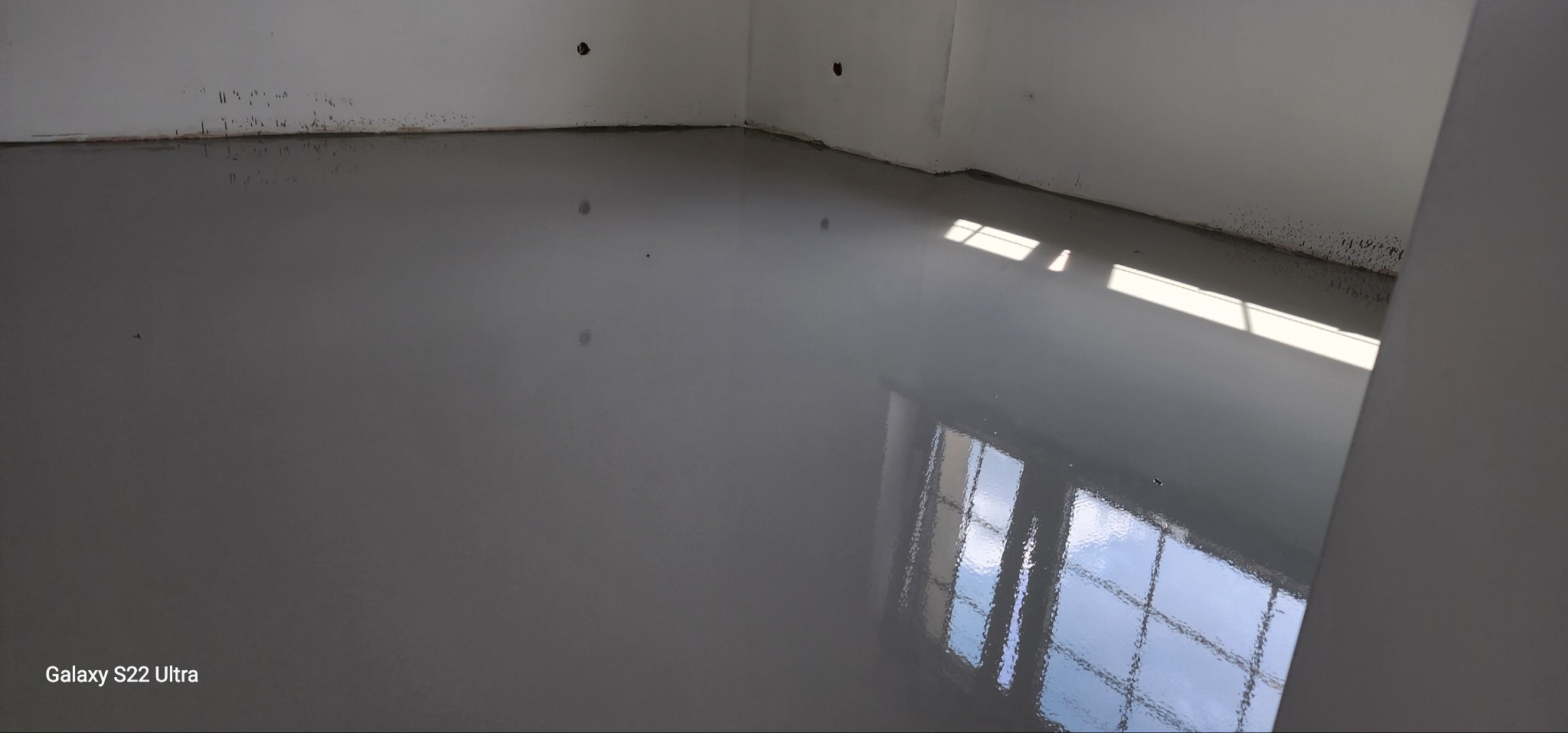 Świeżo wylana, gładka, szara posadzka samopoziomująca w rogu pomieszczenia, z odbiciem okna i smugami światła. Ściany białe, surowe.