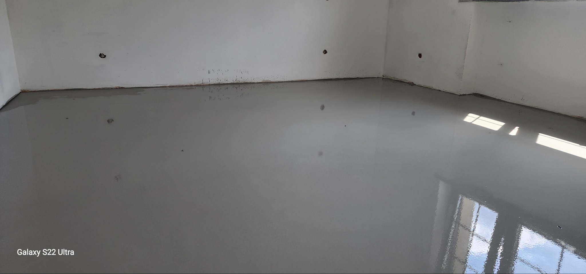 Wylany, samopoziomujący szary podkład podłogowy w rogu pomieszczenia z widocznymi otworami na gniazdka elektryczne w ścianie i odbiciem okna w podłodze.