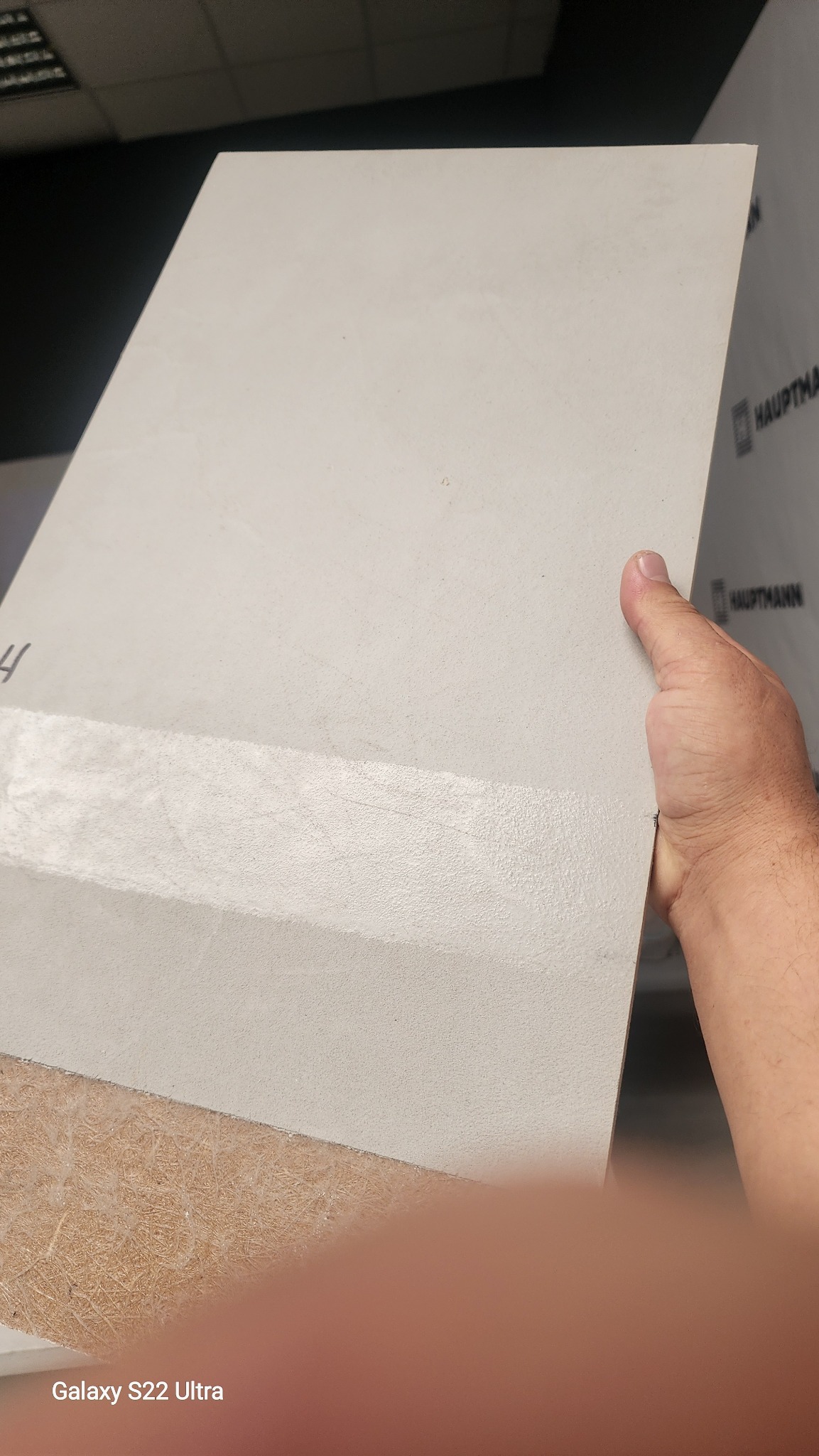 Próbka mikrocementu w dłoni we Wrocławiu. Widoczna struktura i odcień szarości na płycie. Na dole fragment podłoża. Prezentacja materiału wykończeniowego.