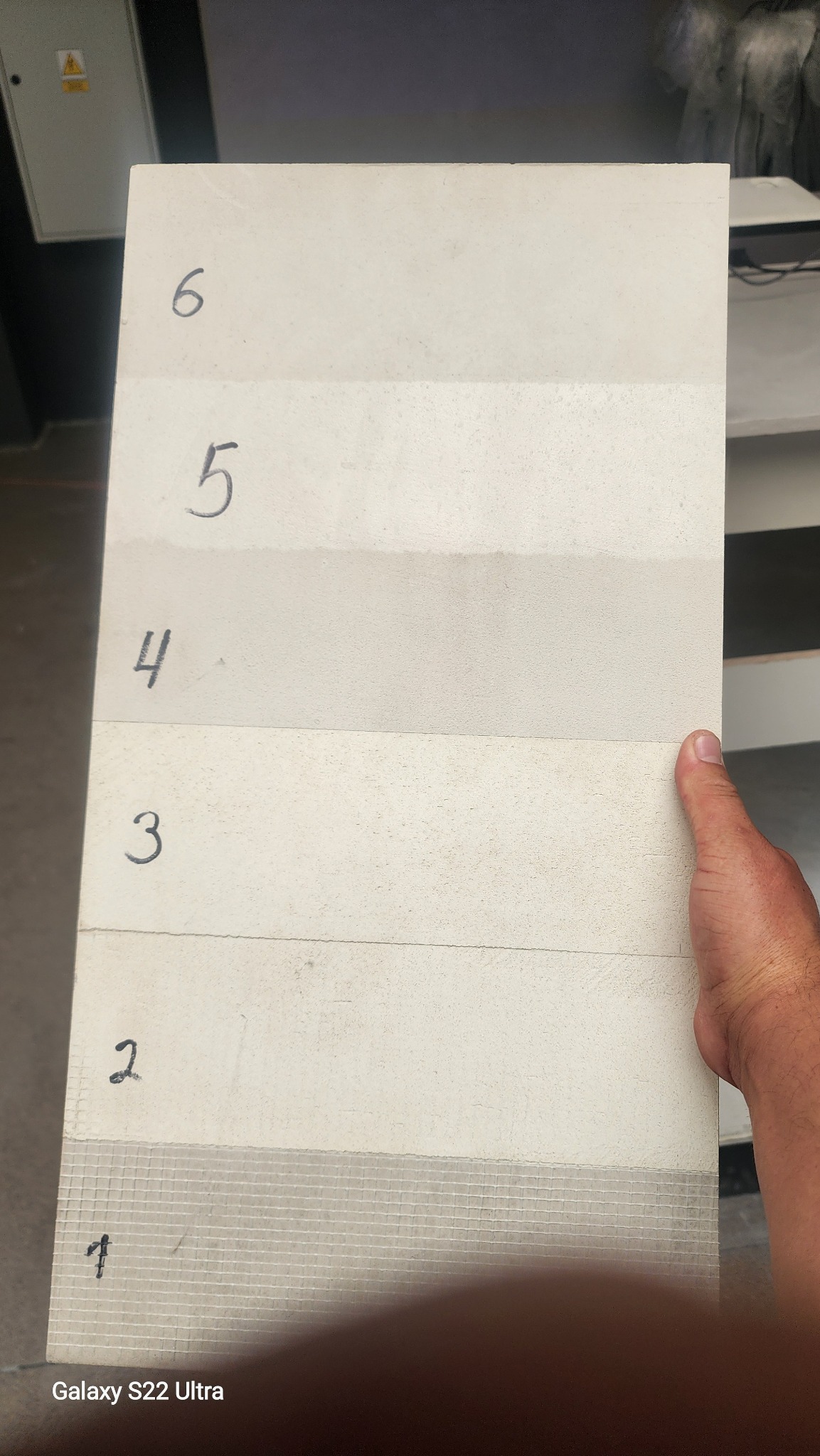 Próbka elewacji z numerami od 1 do 6, ukazująca różne tekstury i odcienie bieli. Widoczna siatka wzmacniająca na próbce nr 1. Ręka trzyma próbkę.