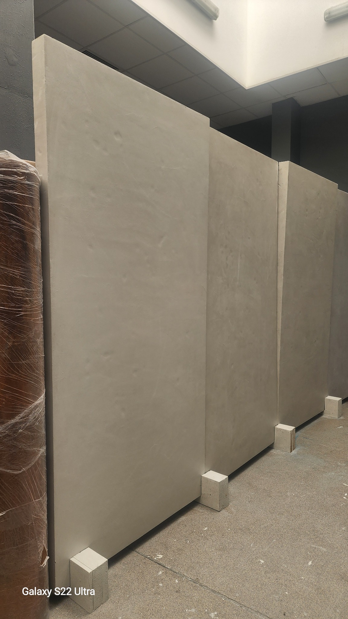 Wykończone ściany w odcieniach beżu, wsparte na białych bloczkach. Widoczna faktura tynku i fragment zabezpieczonego folią materiału budowlanego.