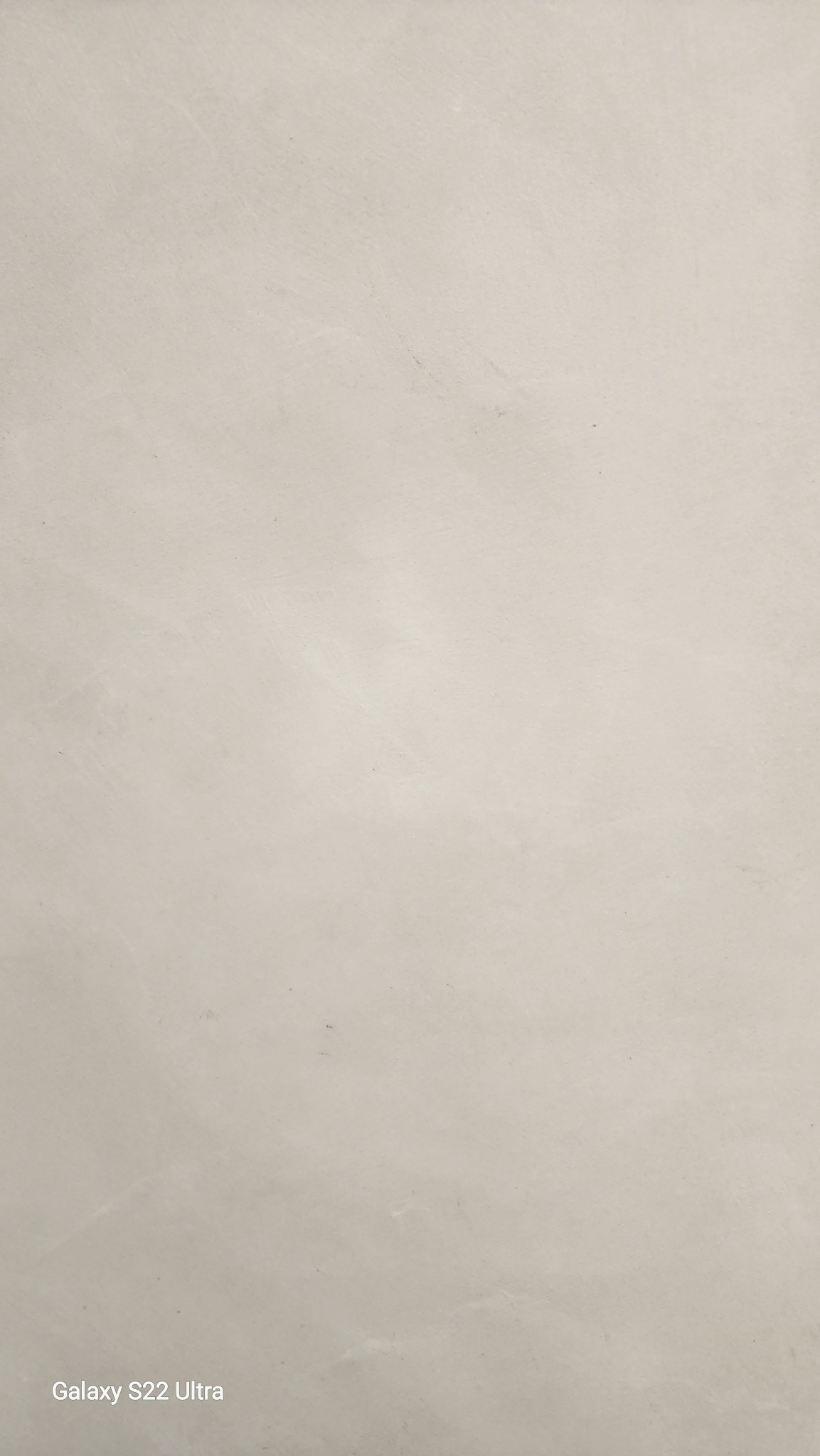 Gładka, beżowa powierzchnia ściany, sfotografowana z bliska, z widoczną subtelną fakturą i delikatnymi smugami światła. Zdjęcie wykonane telefonem Galaxy S22 Ultra.