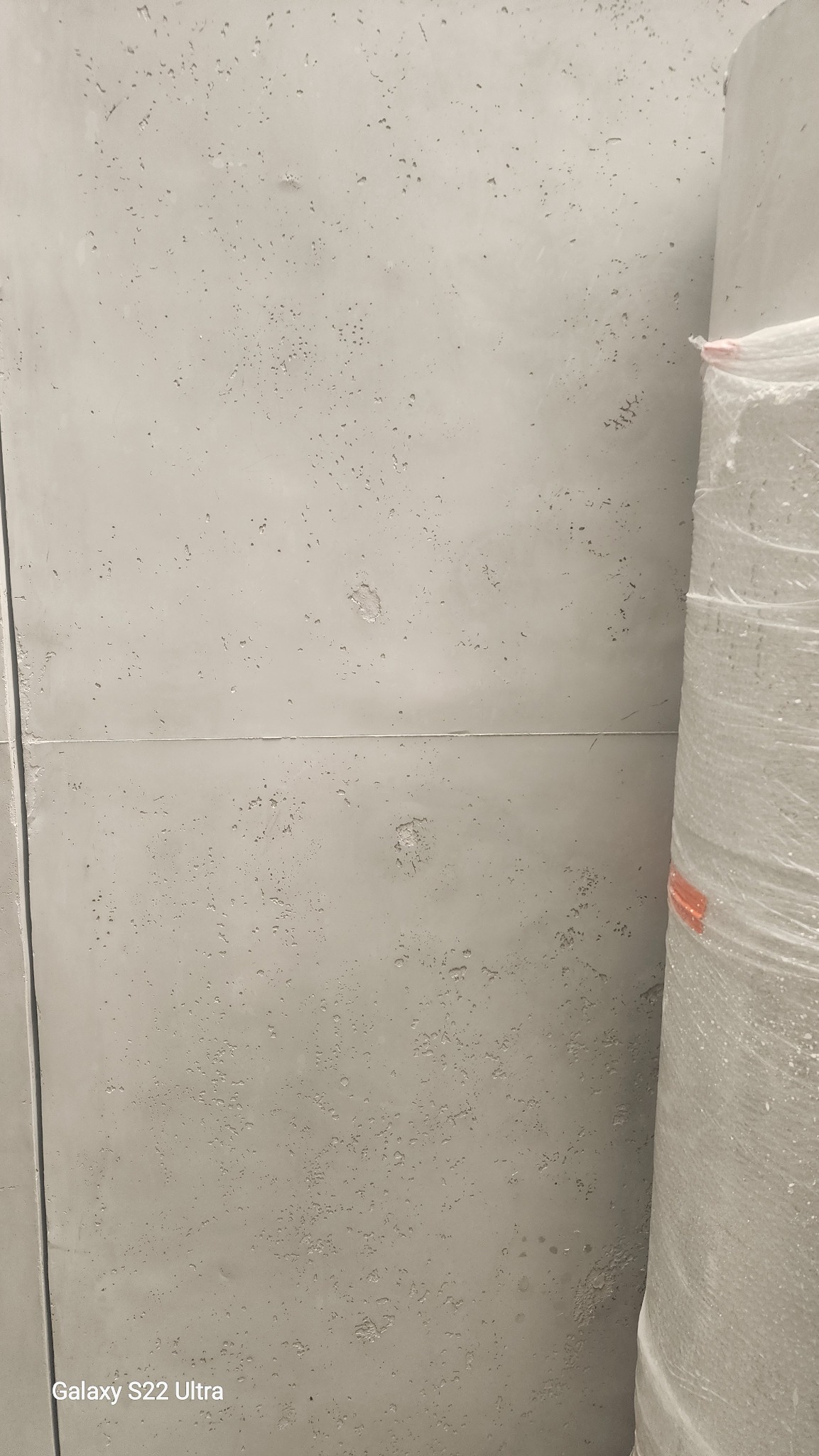 Ściana z betonu architektonicznego z widoczną fakturą i łączeniami płyt. Obok rolka materiału budowlanego w folii. Minimalistyczne wykończenie wnętrza.