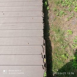 ZAG-BUD USŁUGI WYKOŃCZENIOWE i BUDOWLANE - Szare deski tarasowe kompozytowe, ułożone równolegle, przylegające do trawy; widoczne niedociągnięcia przy krawędzi, z drobnymi roślinami wyrastającymi spod desek.