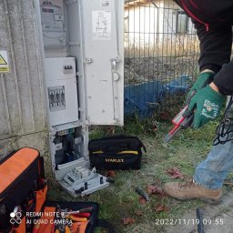 ZAG-BUD USŁUGI WYKOŃCZENIOWE i BUDOWLANE - Elektryk w zielonych rękawicach łączy przewody w otwartej skrzynce elektrycznej na zewnątrz, obok narzędzi i torby Stanley.