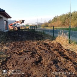 ZAG-BUD USŁUGI WYKOŃCZENIOWE i BUDOWLANE - Czerwony wózek widłowy Manitu z łyżką na nierównym, błotnistym terenie obok częściowo wykończonego budynku i zielonego ogrodzenia siatkowego.