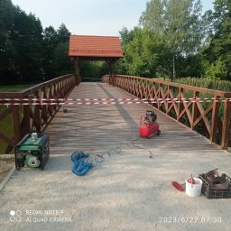 ZAG-BUD USŁUGI WYKOŃCZENIOWE i BUDOWLANE - Drewniany most z czerwonym zadaszeniem, zabezpieczony taśmą ostrzegawczą, z widocznym agregatem prądotwórczym, kompresorem i narzędziami malarskimi, prawdopodobnie w trakcie renowacji.