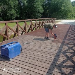 ZAG-BUD USŁUGI WYKOŃCZENIOWE i BUDOWLANE - Mężczyzna w okularach ochronnych maluje drewniany most pistoletem natryskowym; obok stoi niebieski agregat prądotwórczy.