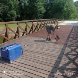 Mężczyzna w okularach ochronnych maluje drewniany most pistoletem natryskowym; obok stoi niebieski agregat prądotwórczy.