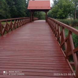 ZAG-BUD USŁUGI WYKOŃCZENIOWE i BUDOWLANE - Drewniany most w kolorze brązowym z zadaszeniem z czerwonej dachówki, prowadzący przez wodę w otoczeniu zieleni.