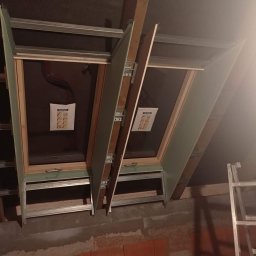 ZAG-BUD USŁUGI WYKOŃCZENIOWE i BUDOWLANE - Montaż dwóch okien dachowych z widocznymi elementami konstrukcyjnymi, profile metalowe, płyty gipsowo-kartonowe i ceglany mur w surowym stanie.