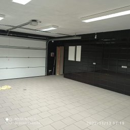 ZAG-BUD USŁUGI WYKOŃCZENIOWE i BUDOWLANE - Wykończone wnętrze garażu z białą bramą segmentową, kremową podłogą z płytek i czarną ścianą z połyskujących płytek, oświetlone lampami sufitowymi.