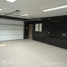 Wykończone wnętrze garażu z białą bramą segmentową, kremową podłogą z płytek i czarną ścianą z połyskujących płytek, oświetlone lampami sufitowymi.