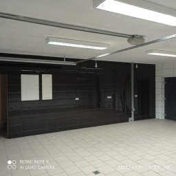 ZAG-BUD USŁUGI WYKOŃCZENIOWE i BUDOWLANE - Wykończone wnętrze pomieszczenia z błyszczącymi, ciemnymi płytkami na ścianach, jasnymi płytkami na podłodze, widocznymi instalacjami elektrycznymi pod sufitem i białymi drzwiczkami rewizyjnymi.