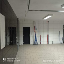 ZAG-BUD USŁUGI WYKOŃCZENIOWE i BUDOWLANE - Wnętrze pomieszczenia z nową, jasną podłogą z płytek i białymi ścianami z widocznymi pionowymi rurami w kolorach czerwonym i niebieskim, czarne błyszczące płytki na ścianie po lewej stronie, czarne...