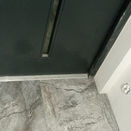 ZAG-BUD USŁUGI WYKOŃCZENIOWE i BUDOWLANE - Ciemne drzwi wejściowe z wąskim przeszkleniem, szara marmurowa podłoga i biała ściana z wystającym gniazdkiem elektrycznym.