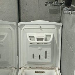 ZAG-BUD USŁUGI WYKOŃCZENIOWE i BUDOWLANE - Wnętrze otwartej pralki automatycznej ładowanej od góry, widoczny bęben i instrukcja dozowania detergentu, na tle szarej ściany z grzejnikiem drabinkowym.