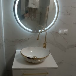 ZAG-BUD USŁUGI WYKOŃCZENIOWE i BUDOWLANE - Biała umywalka nablatowa ze złotym zdobieniem, złota bateria, okrągłe lustro z podświetleniem LED, marmurowe płytki na ścianach, grzejnik w tle odbity w lustrze.