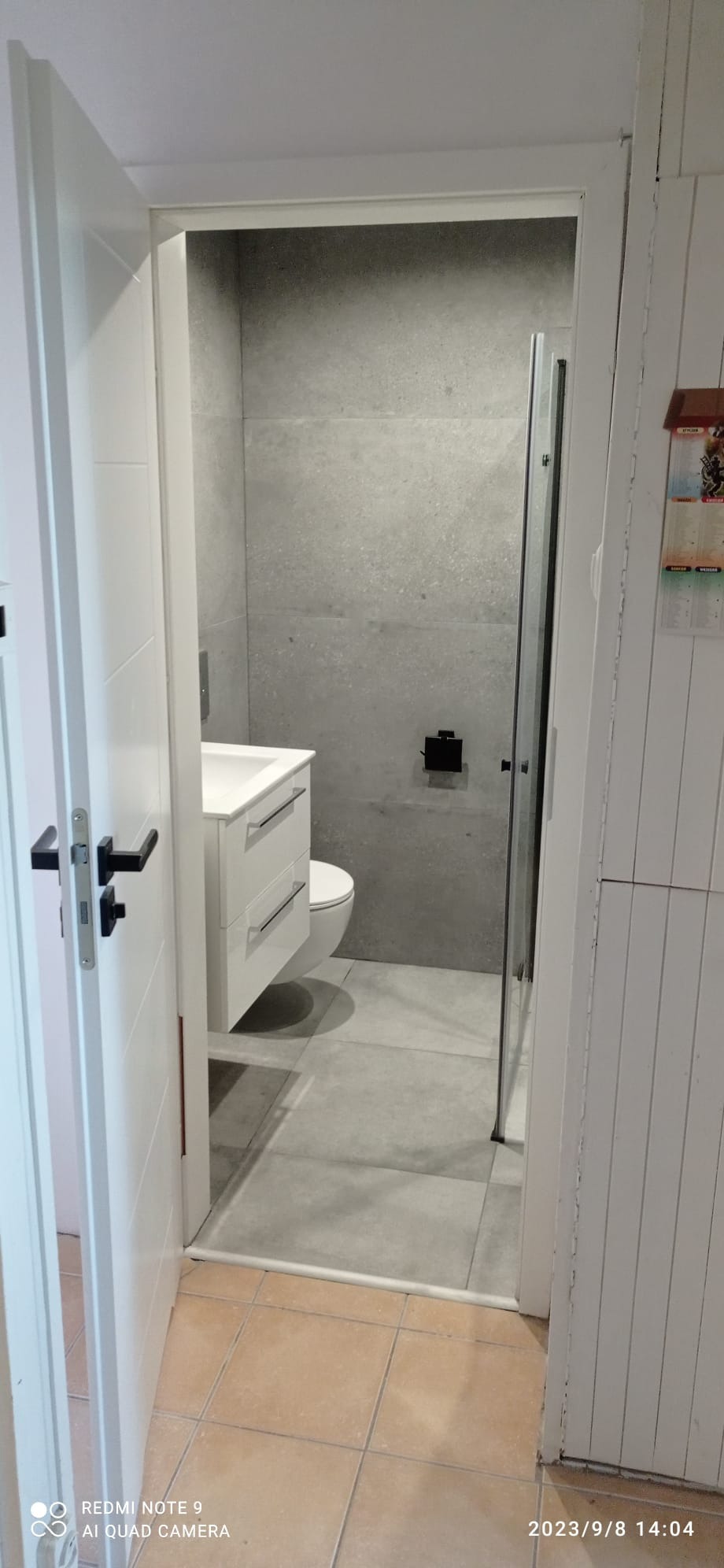 Wykończona łazienka z szarymi płytkami, białą szafką z umywalką i kabiną prysznicową widziana zza otwartych białych drzwi.