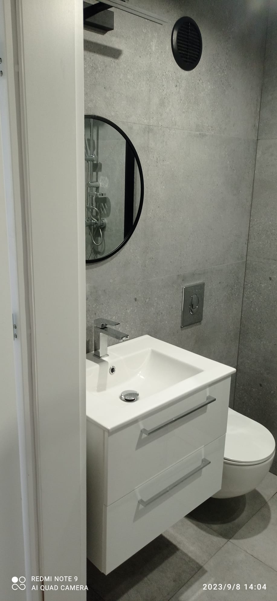 Nowoczesna łazienka w szarościach: biała szafka pod umywalką z chromowaną baterią, okrągłe lustro z czarną ramą odbijające prysznic, wisząca toaleta i szare płytki na ścianie.