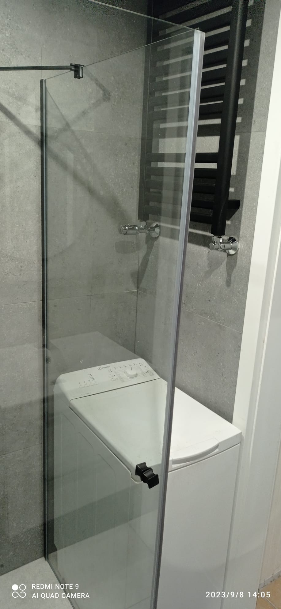 Szklana kabina prysznicowa z czarnymi okuciami, przylegająca do pralki w łazience wykończonej szarymi płytkami.