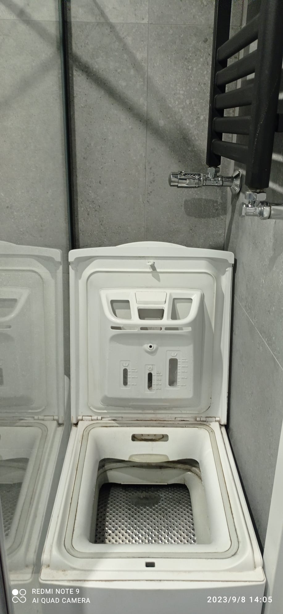 Wnętrze otwartej pralki automatycznej ładowanej od góry, widoczny bęben i instrukcja dozowania detergentu, na tle szarej ściany z grzejnikiem drabinkowym.