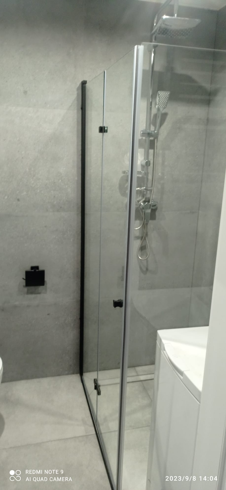 Nowoczesna kabina prysznicowa z czarnymi profilami, szklane drzwi, deszczownica i bateria w chromie, szare płytki na ścianach, czarny uchwyt na papier toaletowy, fragment pralki.