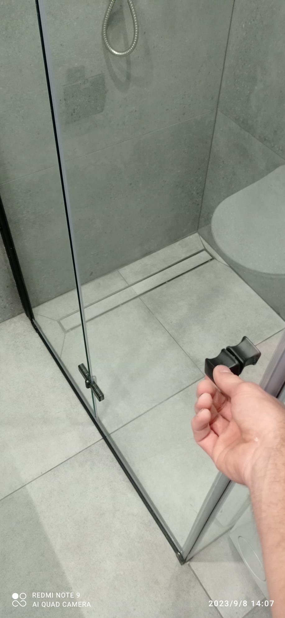 Szklana kabina prysznicowa z czarnymi profilami, uchwyt otwierany przez dłoń, szare płytki podłogowe i ścienne, odpływ liniowy przy ścianie.