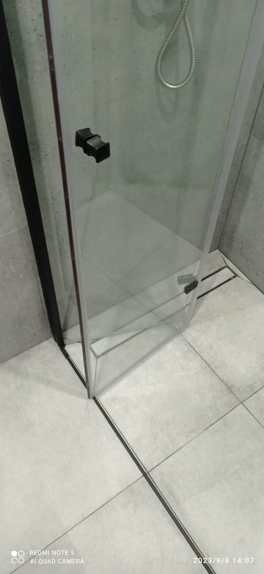 Szklana kabina prysznicowa z czarnymi detalami, zamontowana w rogu łazienki wyłożonej szarymi płytkami, widoczny odpływ liniowy w podłodze.