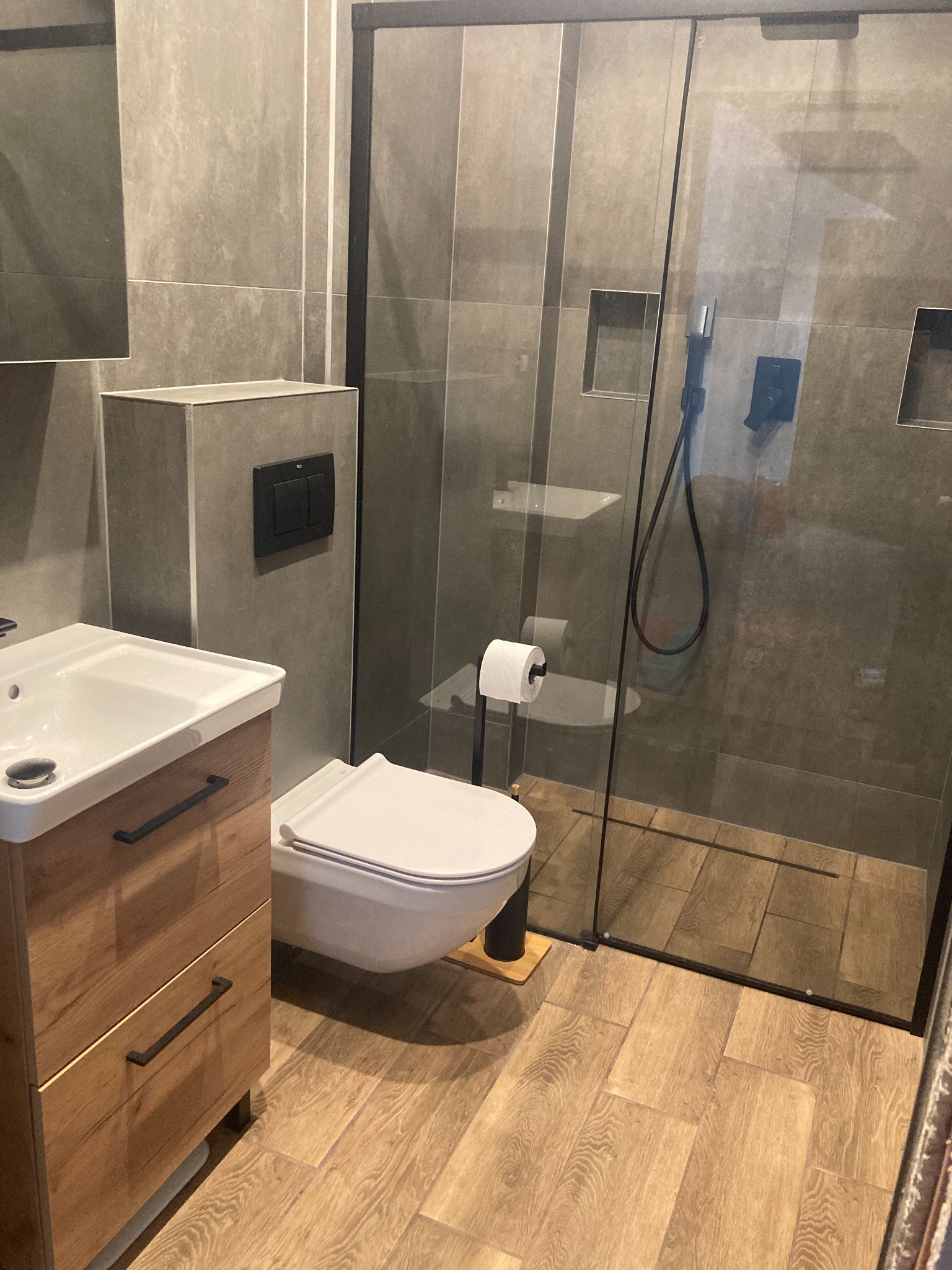 Nowoczesna łazienka z szarą glazurą, drewnopodobną podłogą, wiszącą toaletą i kabiną prysznicową ze szklanymi drzwiami, widoczna umywalka z szafką z drewnianym frontem.