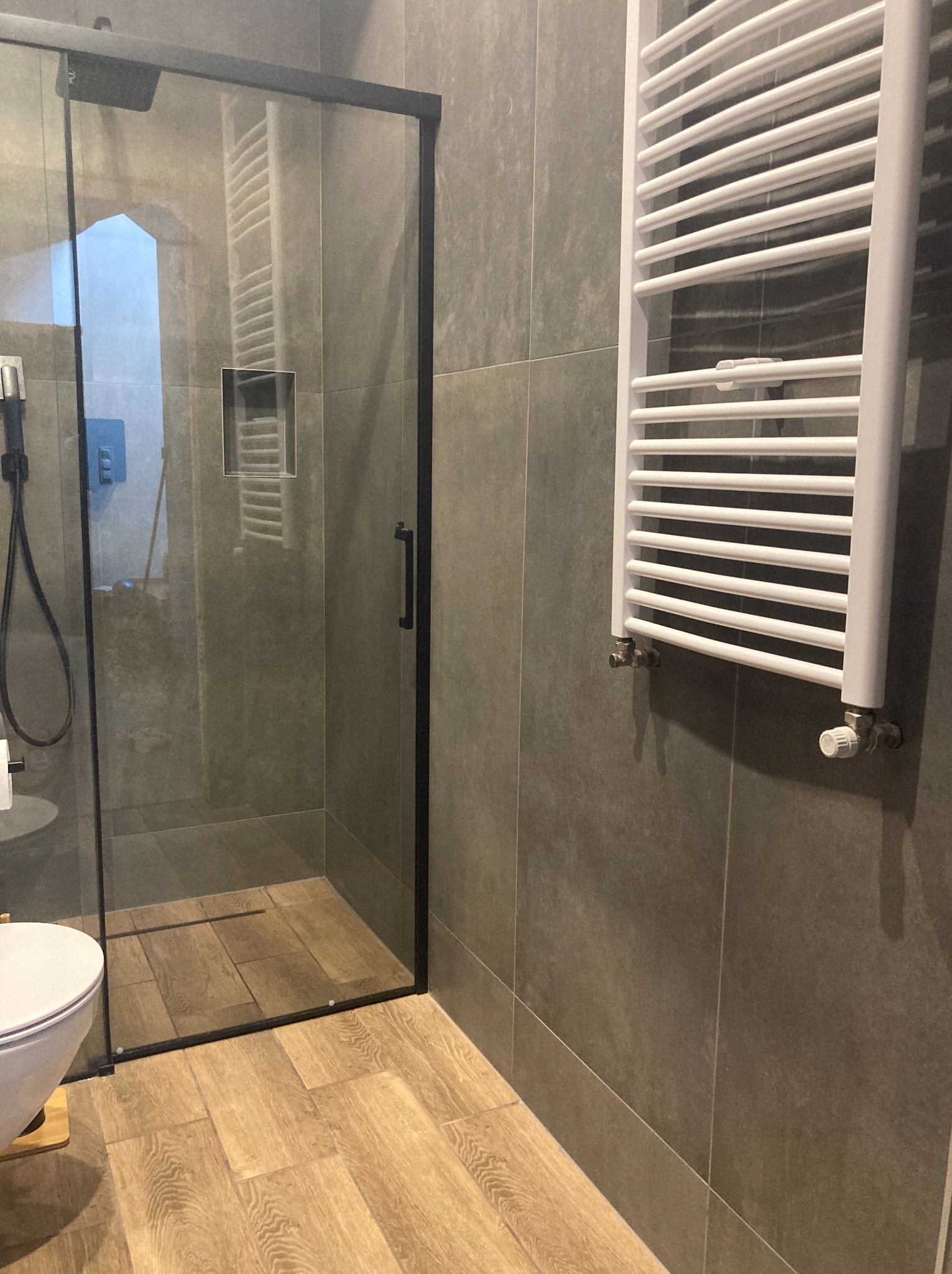 Szara łazienka z kabiną prysznicową w czarnej ramie, drewnopodobną podłogą i białym grzejnikiem drabinkowym na ścianie z szarych płytek.