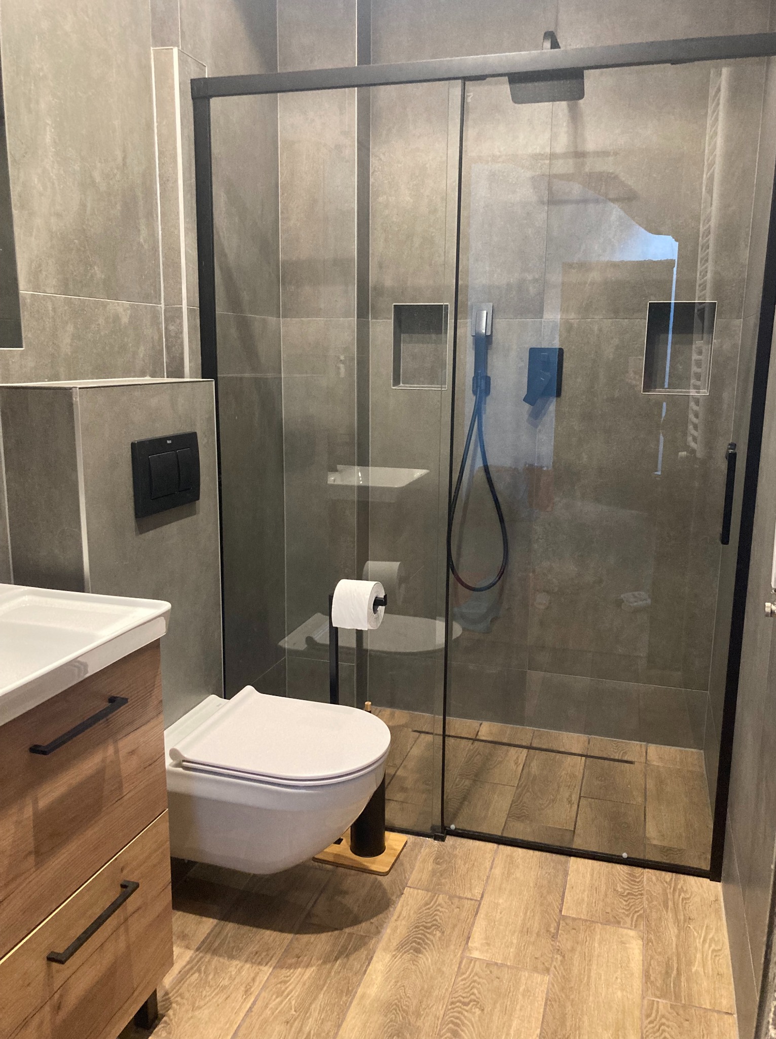 Nowoczesna łazienka z szarą glazurą, kabiną prysznicową typu walk-in z czarnymi profilami, podwieszaną toaletą i drewnianą szafką pod umywalką z czarnymi uchwytami.