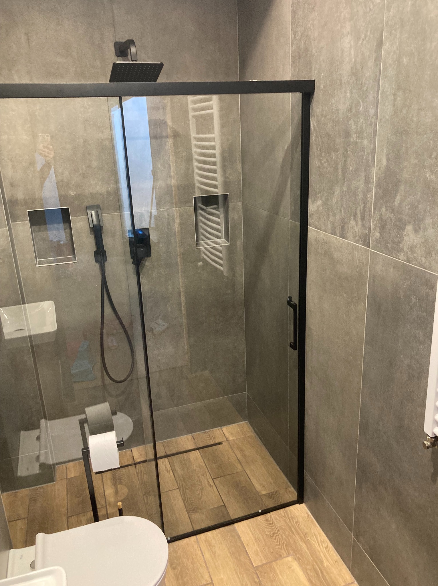 Kabina prysznicowa z czarnymi profilami, szara glazura na ścianach i drewnopodobna podłoga, widoczna toaleta i akcesoria łazienkowe wewnątrz.