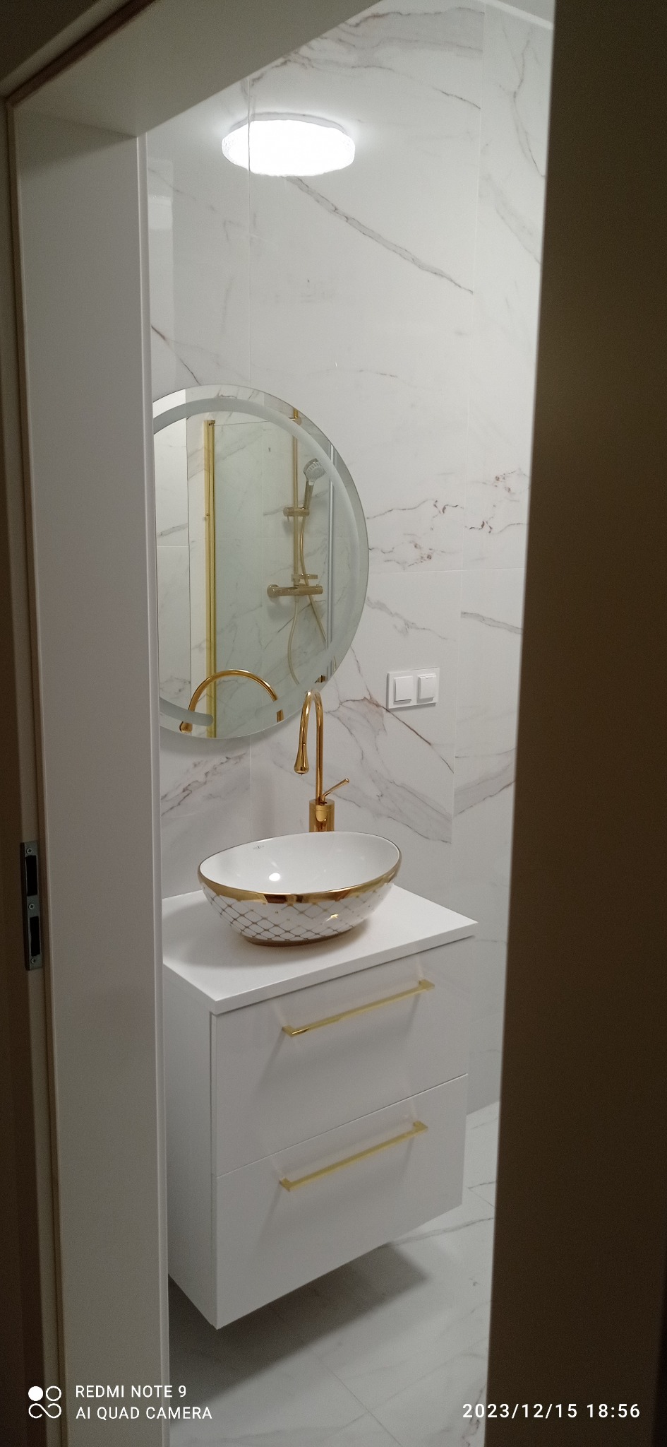 Nowoczesna łazienka z umywalką nablatową w geometryczny wzór z złotymi akcentami, złotą armaturą i okrągłym lustrem z podświetleniem, marmurowe płytki na ścianach, minimalistyczny design.