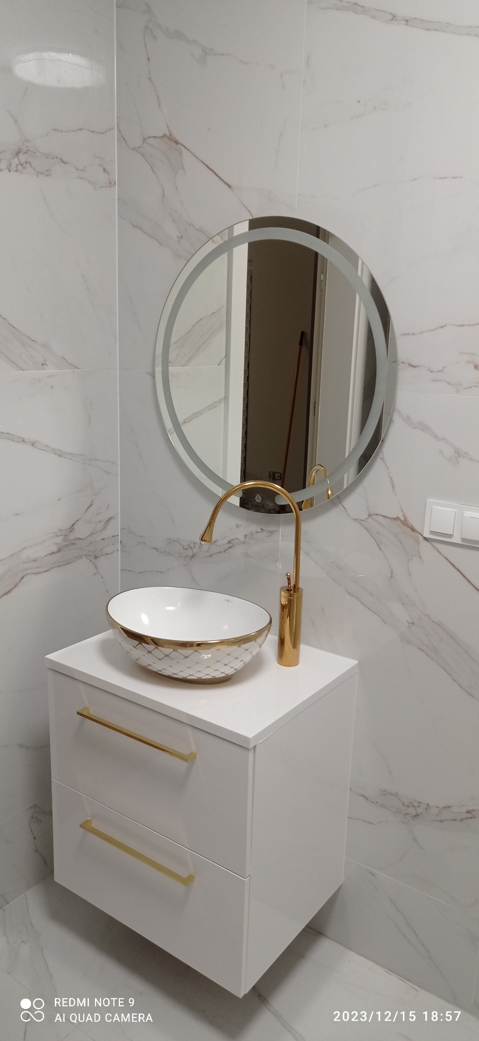Biała szafka łazienkowa z umywalką nablatową w złote wzory, bateria w kolorze złota i okrągłe lustro z podświetleniem LED na ścianie wyłożonej marmurowymi płytkami.