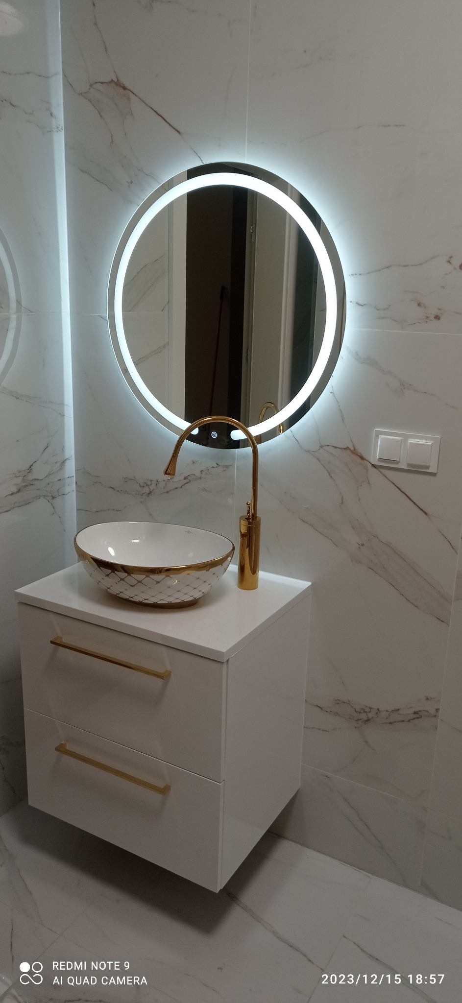 Biała szafka łazienkowa z dwiema szufladami i złotymi uchwytami, na niej umywalka nablatowa z dekoracyjnym złotym wzorem i złotą armaturą, w tle okrągłe lustro z podświetleniem LED oraz marmurowe...