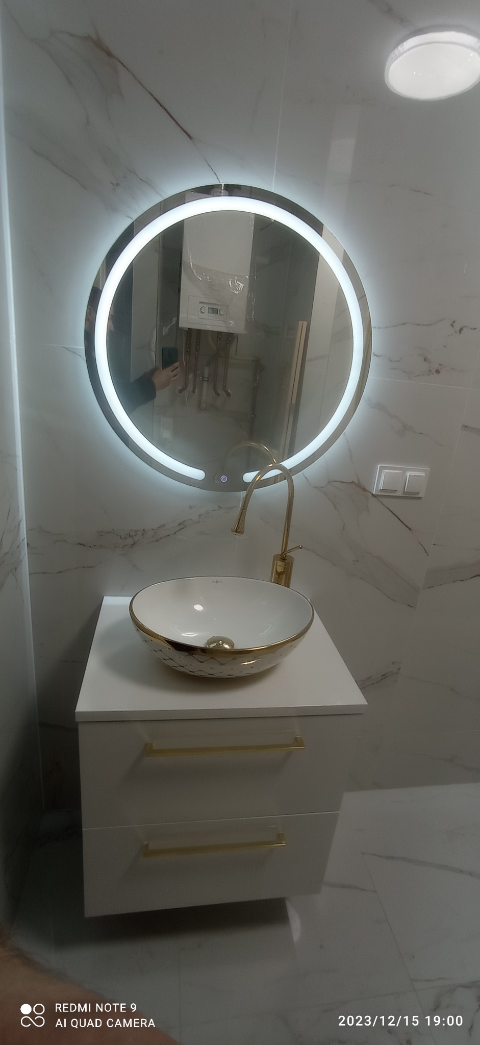 Biała umywalka nablatowa ze złotym zdobieniem, złota bateria, okrągłe lustro z podświetleniem LED, marmurowe płytki na ścianach, grzejnik w tle odbity w lustrze.