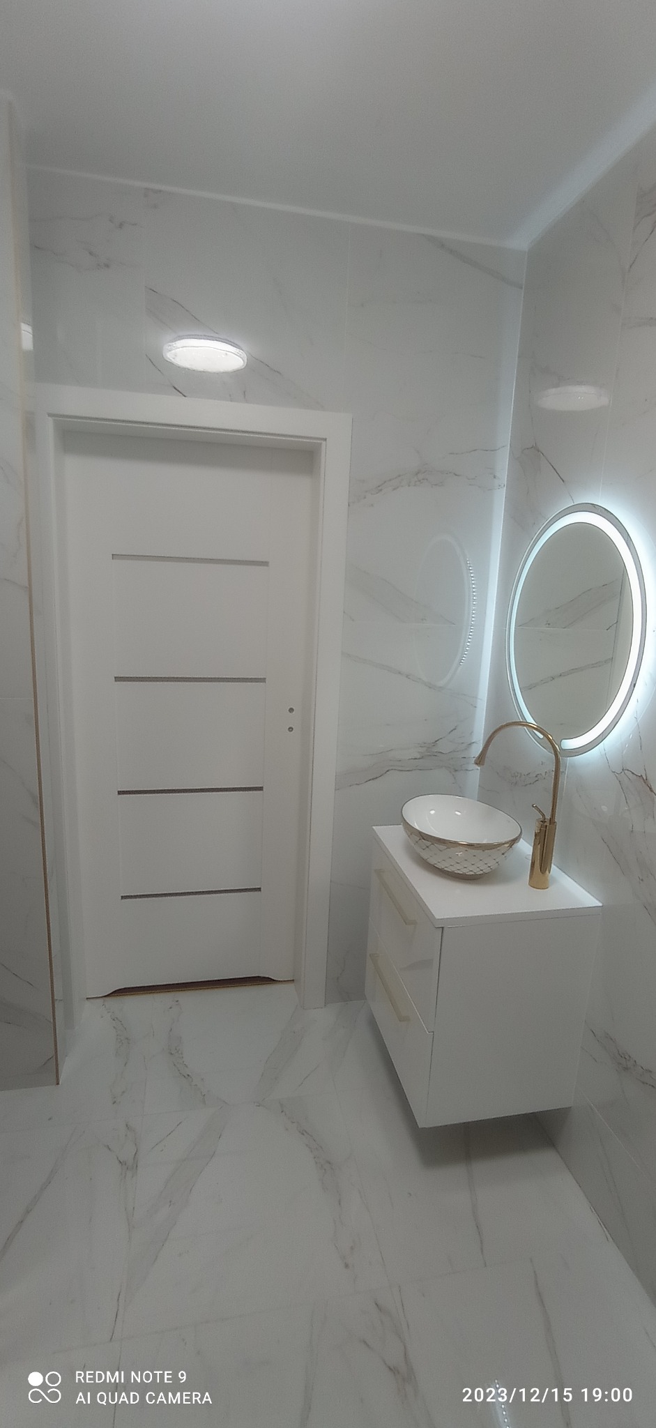 Nowoczesna łazienka z białymi marmurowymi płytkami, białą szafką z umywalką w stylu glamour z pozłacaną armaturą i okrągłym lustrem z podświetleniem LED, obok białe drzwi z poziomymi frezami.