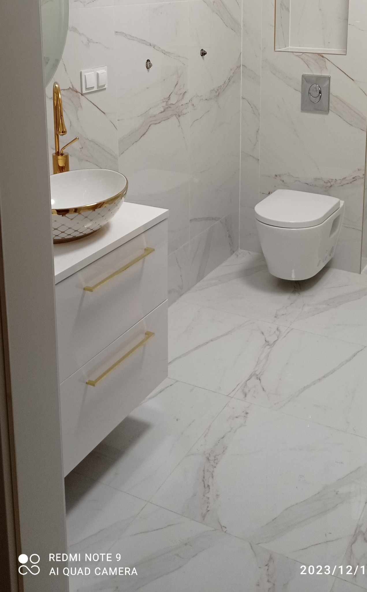 Nowoczesna łazienka z marmurowymi płytkami, wiszącą toaletą i umywalką nablatową z baterią w kolorze złotym oraz szafką z białym frontem i złotymi uchwytami.