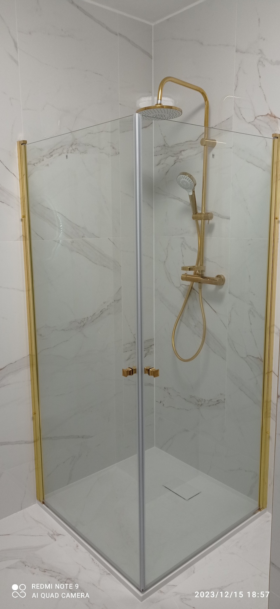 Narożna kabina prysznicowa z brodzikiem, złote profile i armatura na tle marmurowych płytek ściennych.