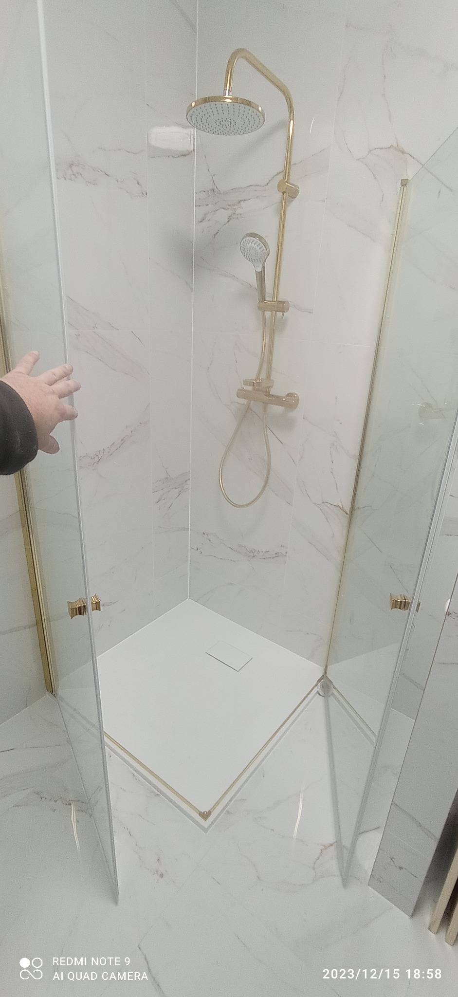 Nowoczesna kabina prysznicowa z białym brodzikiem i złotą armaturą na tle marmurowych płytek; widoczna dłoń przy szklanych drzwiach.