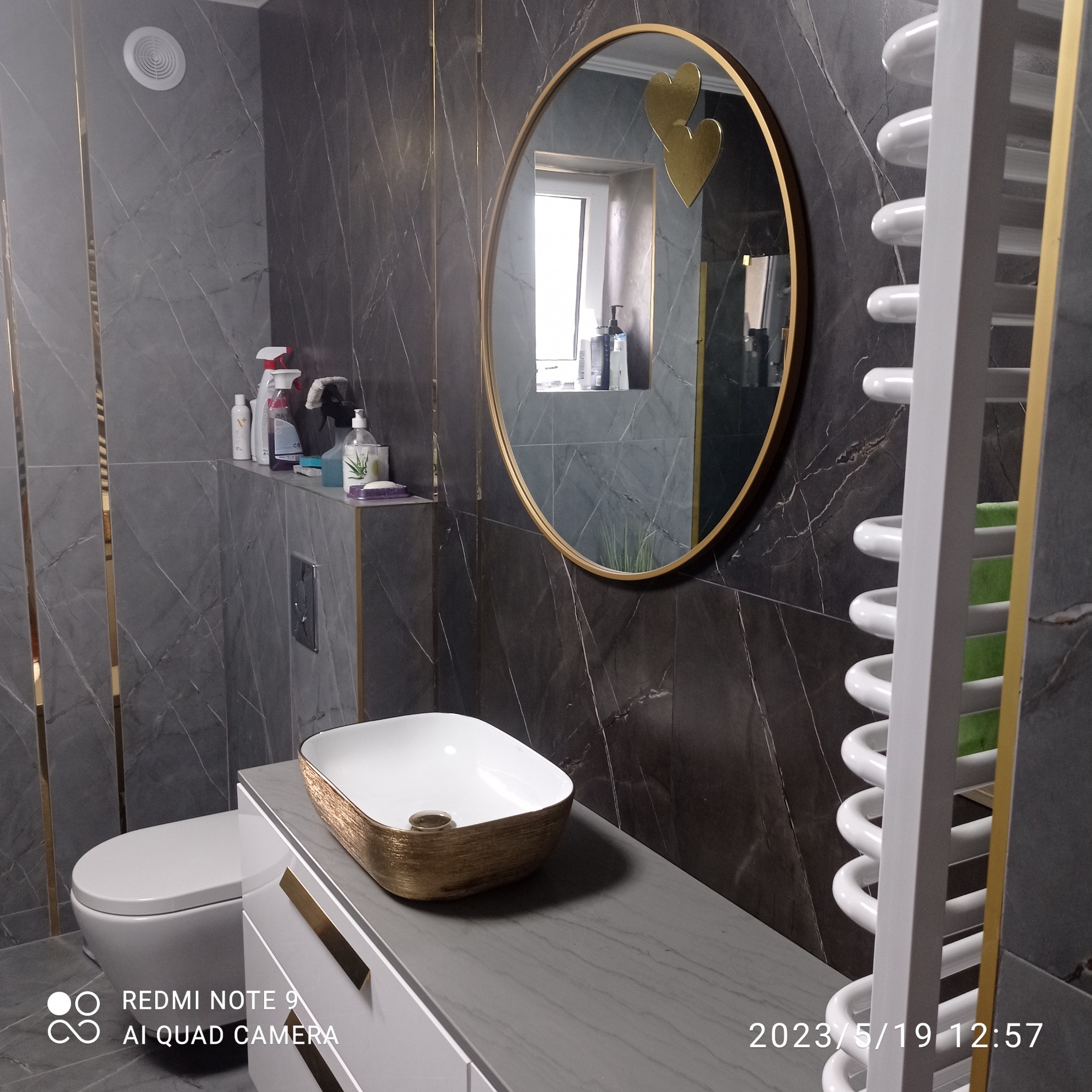 Elegancka łazienka z szarymi marmurkowymi płytkami, owalnym lustrem w złotej ramie z dekoracjami w kształcie serc, designerską umywalką ze złotym wykończeniem i białym grzejnikiem drabinkowym.