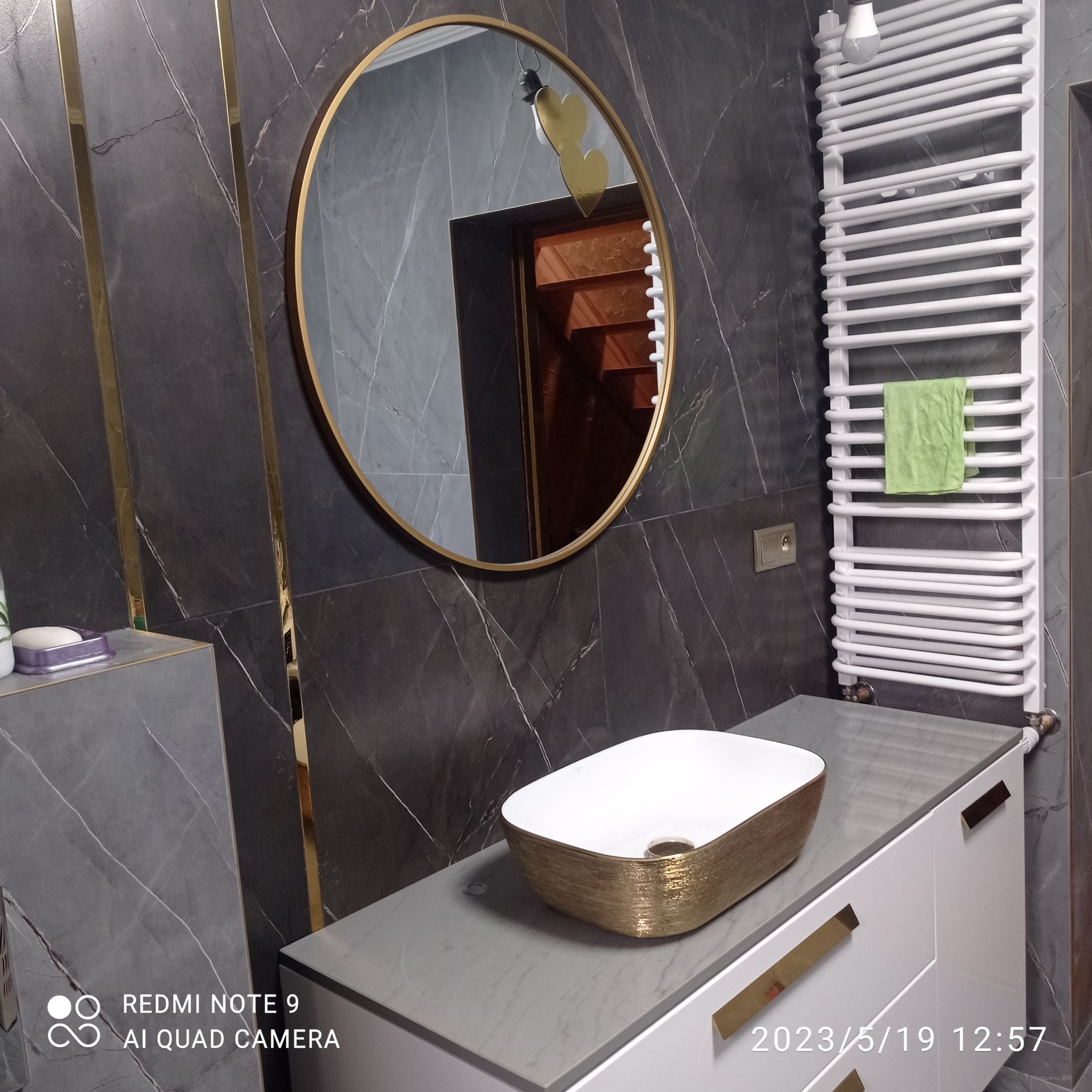 Nowoczesna łazienka z umywalką nablatową w kolorze białym i złotym, okrągłym lustrem z złotą ramą, grzejnikiem drabinkowym i szarymi płytkami z złotymi wstawkami.