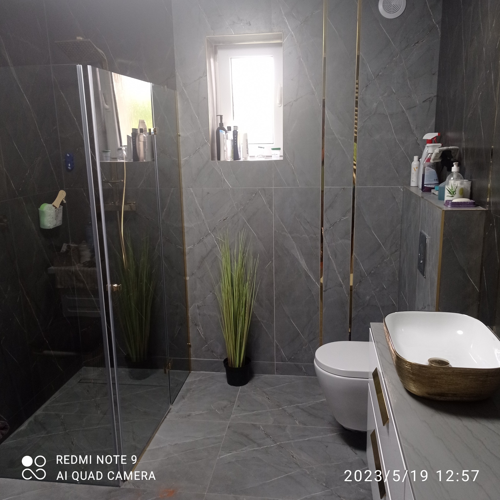 Nowoczesna łazienka z szarymi marmurowymi płytkami, kabiną prysznicową typu walk-in, toaletą wiszącą i umywalką nablatową z złotymi akcentami.