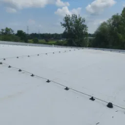 Biały dach płaski z zamontowanymi odgromnikami i elementami wentylacji, widok na okoliczne drzewa i słupy wysokiego napięcia w tle.