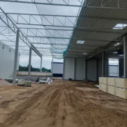 Wnętrze hali w trakcie budowy z widoczną konstrukcją stalową dachu, częściowo pokrytą blachą trapezową i siatką zabezpieczającą, ślady opon na ziemi, przygotowane płyty warstwowe i materiały...