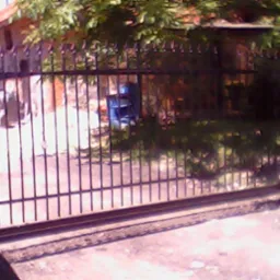 Ciemna, metalowa brama przesuwna z pionowymi prętami na tle zarośniętego ogrodu i budynku z cegły.